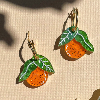 Pair of earrings shaped like oranges with green leaves on a beige background